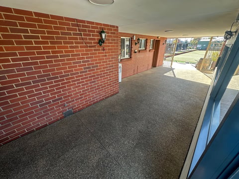 Empty brick room with gray carpet floor, outdoor entrance visible, and windows overlooking green area