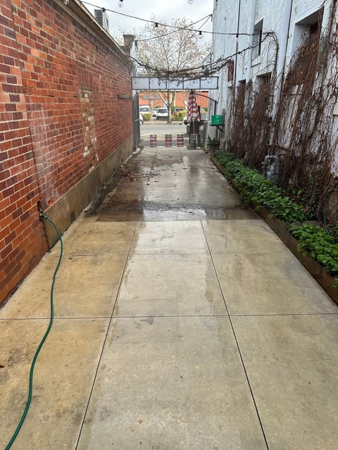 Narrow urban alley with red brick wall on left, weathered concrete pavement, green hose, and buildings at far end